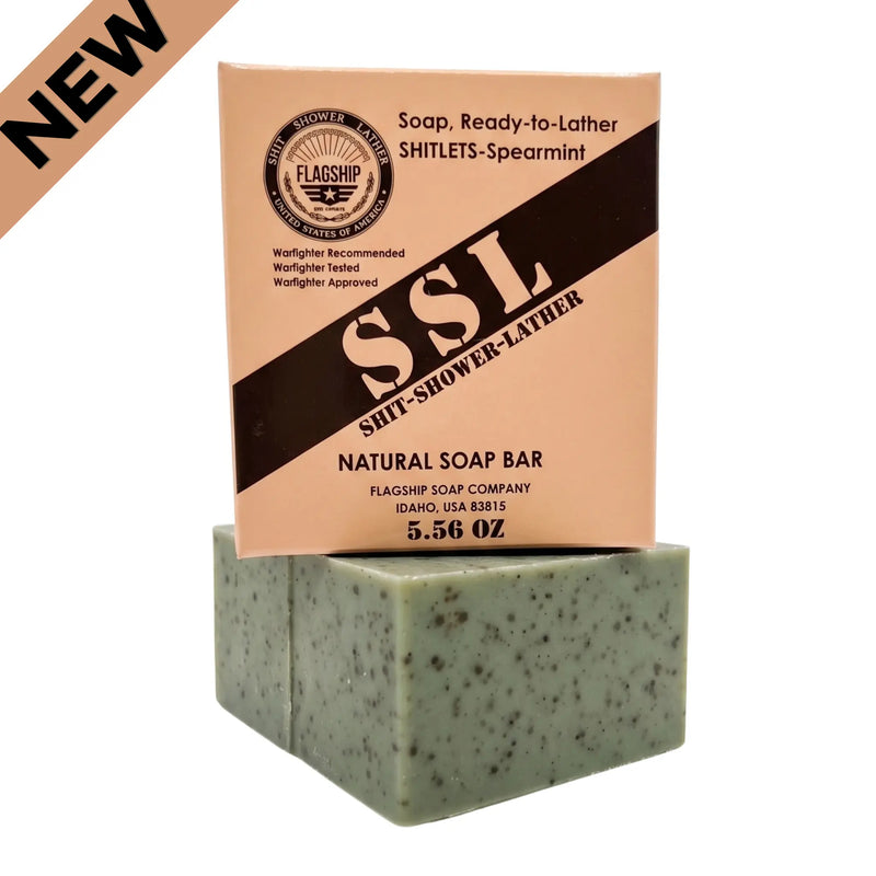 Vertical product shot of 'Flagship Soap Company' natural exfoliating bar (green with coffee specks) stacked beneath its peach-colored cardboard box on a white background. The box displays the 'Flagship' logo, 'NATURAL SOAP', and 'MRE-THEMED GIFT SOAP' text. The underlying soap bar's texture is clearly visible, showcasing the coffee and sea clay exfoliating particles. High-performance, patriotic veteran gift soap from Flagship Soap Company.  New Release