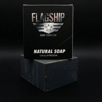 Front view of Flagship Soap Company’s signature "Natural Soap" black box, labeled as 5.56 oz of Freedom. The box is stacked on a thick, artisan-cut activated charcoal soap bar against a clean black background.
