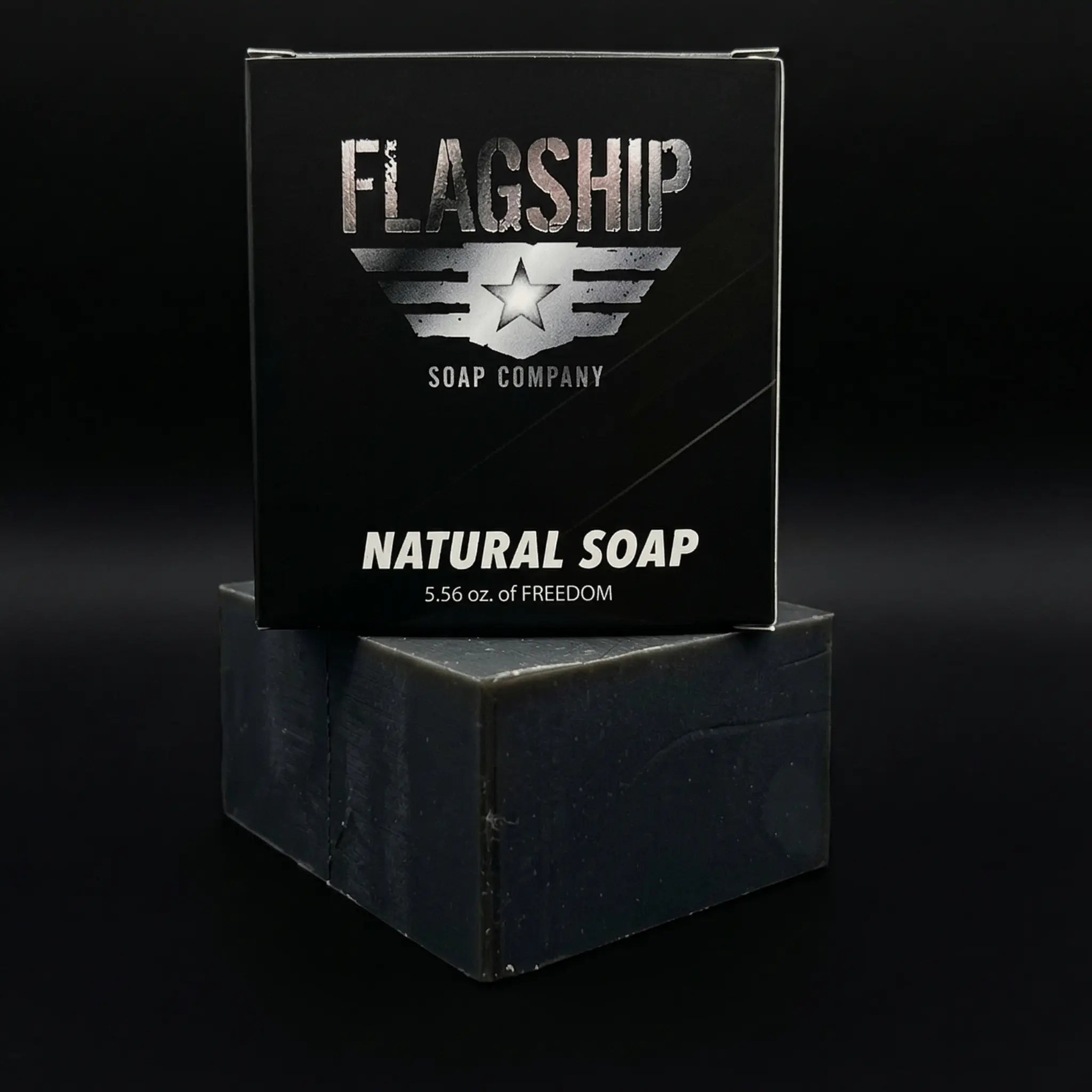 Front view of Flagship Soap Company’s signature "Natural Soap" black box, labeled as 5.56 oz of Freedom. The box is stacked on a thick, artisan-cut activated charcoal soap bar against a clean black background.