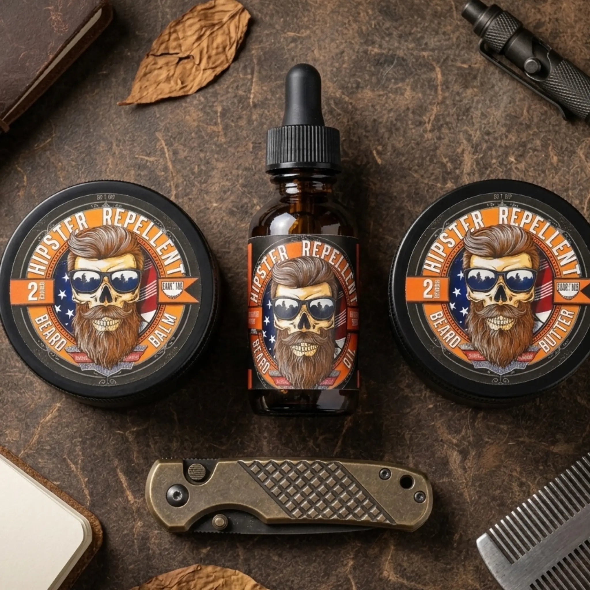 An overhead grouping of Flagship Soap Company's Hipster Repellent 2oz Beard Balm tin, 1oz Beard Oil dropper bottle, and 2oz Beard Butter tin. The rugged bearded skull logo is central, flanked by a tactical folding knife, a metal beard comb, and dried leaves on a weathered leather surface, visually representing the tough yet refined nature of the citrus and cedarwood scented products.