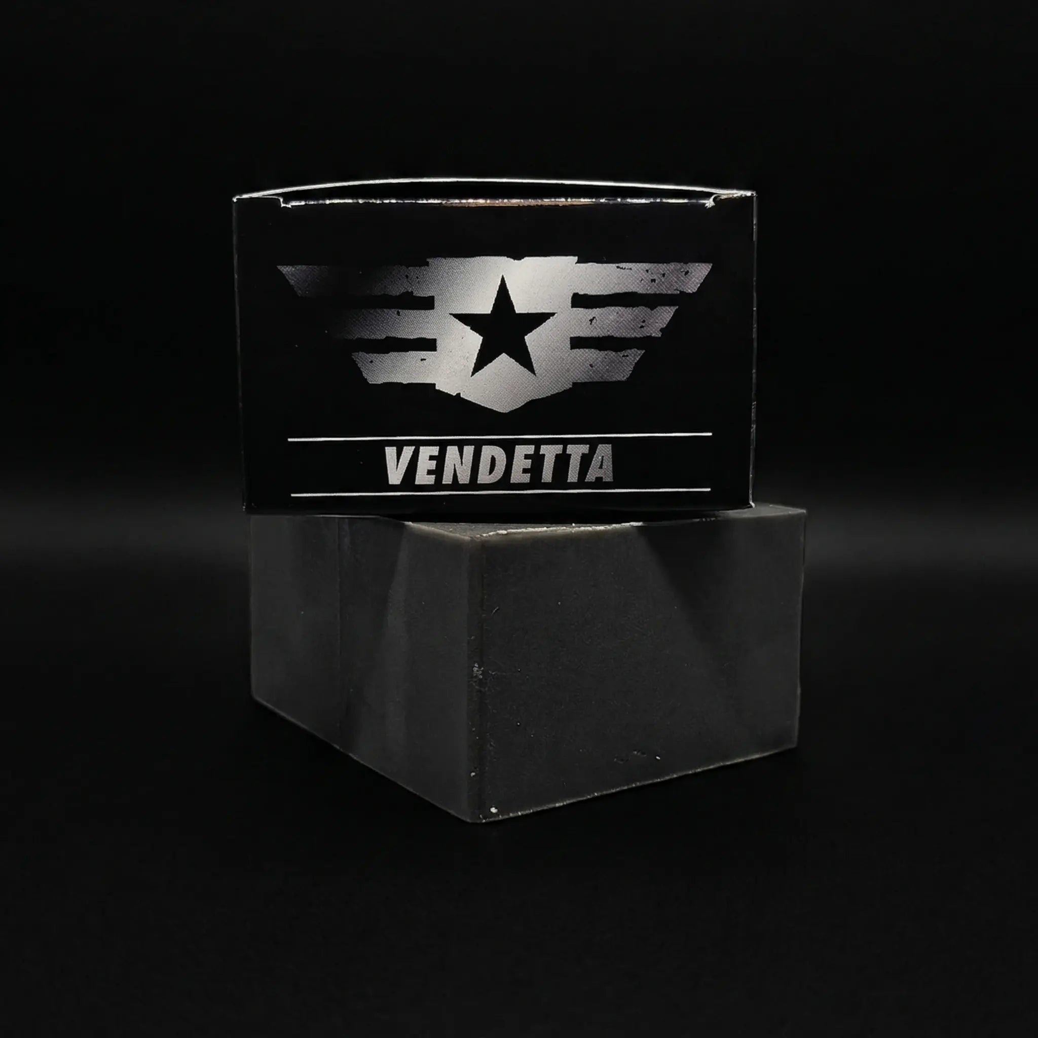 Top view of the Vendetta soap box by Flagship Soap Company resting on a black activated charcoal soap bar. The packaging features a silver tactical wing and star logo against a sleek black box.