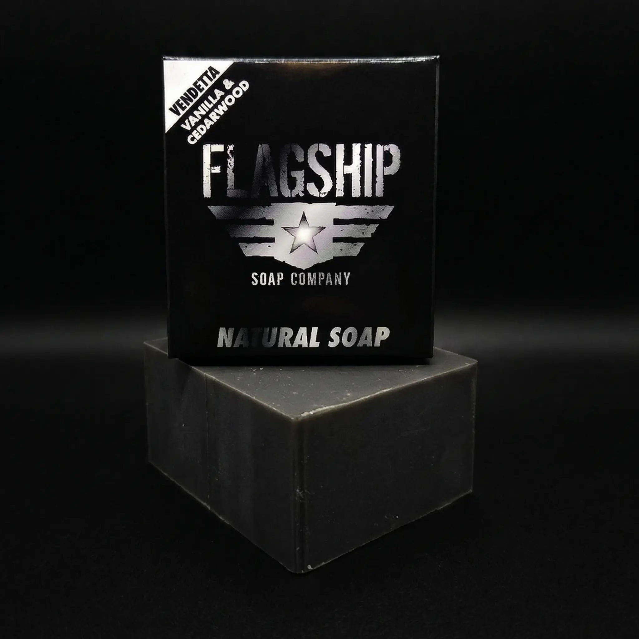 Front-facing shot of the Vendetta soap box by Flagship Soap Company. The corner label identifies the sweet vanilla and cedarwood scent. The box sits atop a rugged, deep-grey activated charcoal soap block.