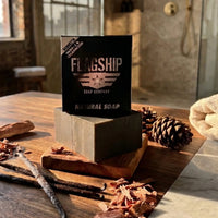 A bar of Vendetta natural soap by Flagship Soap Company on a rustic wooden tray. The scene features sweet vanilla beans, cedarwood shavings, and pinecones in a sunlit, modern bathroom to highlight the rugged scent profile.